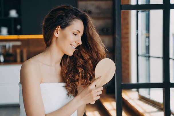 Νεαρή όμορφη ισπανόφωνη γυναίκα βουρτσίζει μακριά υγιή καστανά σγουρά μαλλιά με ξύλινη βούρτσα μαλλιών μετά το ντους. Γυναίκα τυλιγμένη σε πετσέτα κάνει πρωινή ρουτίνα ομορφιάς στο σπίτι. Διαφήμιση καλλυντικών περιποίησης μαλλιών. - Φωτογραφία, εικόνα