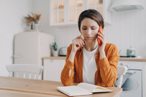 Nervous frowning woman making phone call customer service, having unpleasant conversation, sitting at table with notebook in kitchen. Businesswoman freelancer working at home, discussing project. - Photo, image