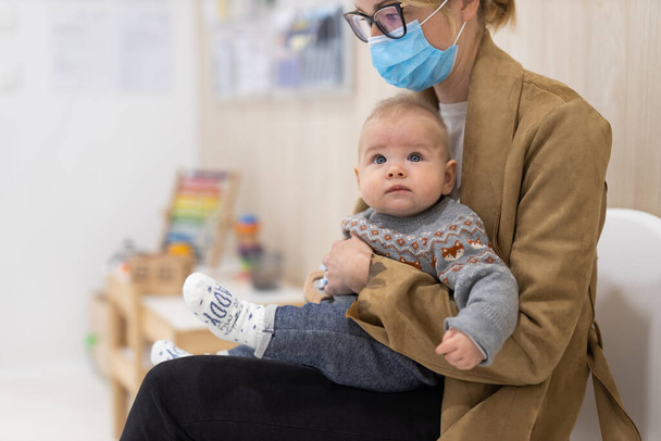 Moeder houdt baby jongen in haar schoot, zittend en wachtend voor het dokterskantoor voor pediatrische putcontrole. kindergezondheidszorgconcept. - Foto, afbeelding