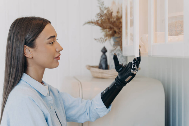 Pretty disabled girl opening kitchen cupboard by bionic prosthetic arm. Young caucasian woman with artificial arm doing chores at home. Advertising of modern prosthesis. Domestic life and disability. - Фото, зображення