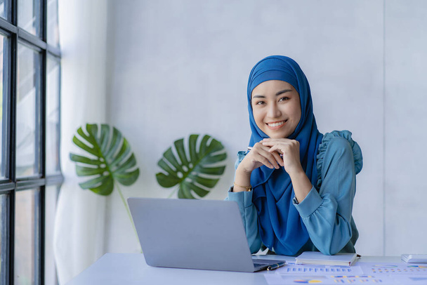 美しいアジア系イスラム教徒の女性は、自宅のリビングルームで財務会計作業を行うために彼女のラップトップを使用してスカーフを身に着けています. - 写真・画像