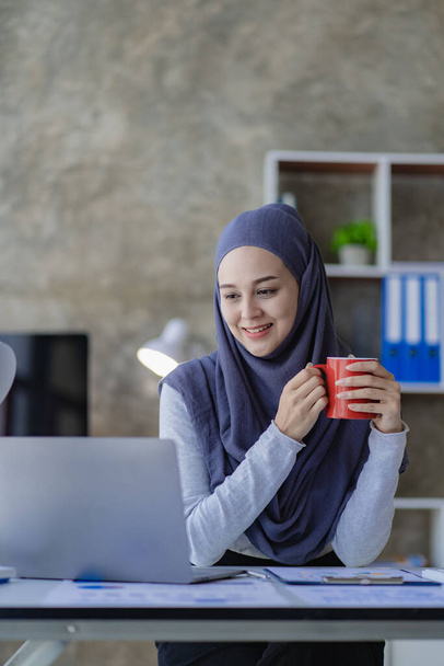 Kaunis aasialainen musliminainen pukeutuu huivi käyttäen hänen kannettava tietokone tehdä taloudellista kirjanpitoa työtä hänen olohuoneessa kotona. - Valokuva, kuva