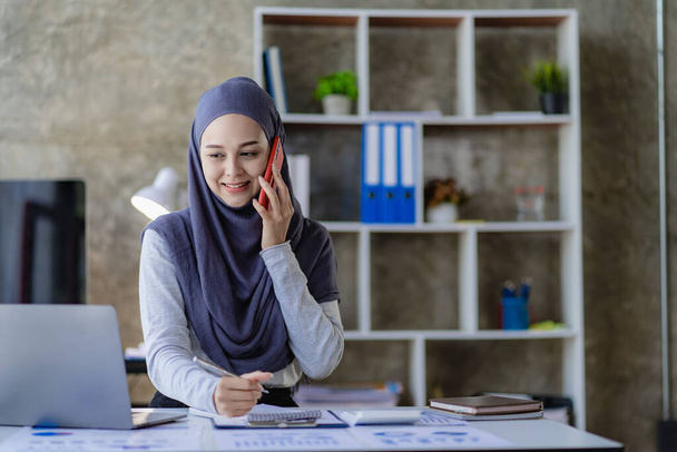美しいアジア系イスラム教徒の女性は、自宅のリビングルームで会計作業を行うためにスマートフォンやノートパソコンを使用してスカーフを身に着けています. - 写真・画像