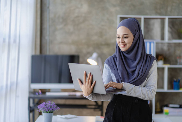 Egy gyönyörű ázsiai muszlim nő fejkendőt visel a laptopjával, hogy pénzügyi könyvelést végezzen otthon a nappalijában.. - Fotó, kép