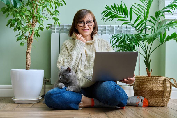 Middle-aged woman in warm clothes with cat using laptop. Female warming at home near heating radiator, with pet in her arms. Cold season winter autumn, leisure lifestyle work freelance, people animals - Φωτογραφία, εικόνα