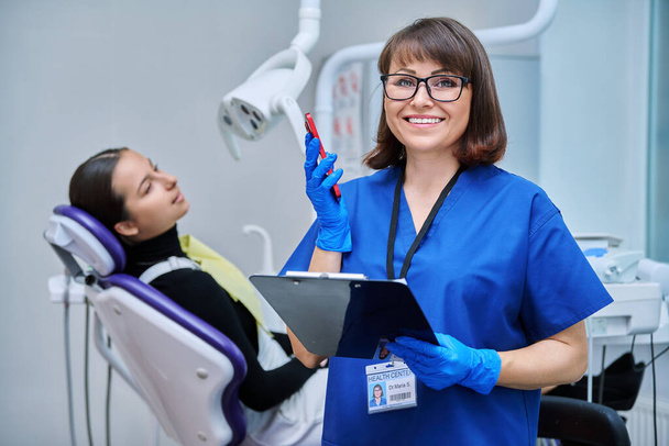 Smiling female dentist, nurse doctor with clipboard phone looking at camera in dental office. Dentistry, hygiene, treatment, medicine, dental health care concept - 写真・画像