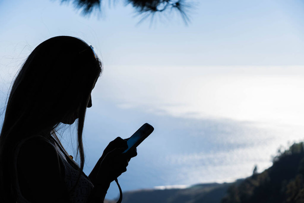 Sylwetka kobiety z długimi włosami i okularami przeciwsłonecznymi w przyrodzie, piszącej za pomocą smartfona nad morzem - Zdjęcie, obraz