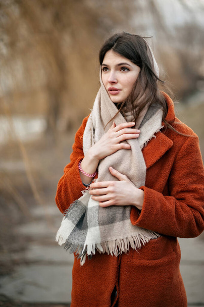 Menina morena jovem, em casaco laranja, olha para o lado com olhar encantado, segurando lenço de lã com as mãos, de pé no parque de outono contra o pano de fundo de árvores embaçadas. - Foto, Imagem
