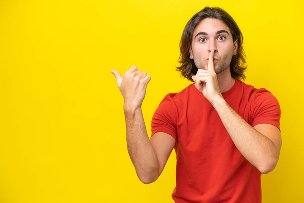 Caucasian handsome man isolated on yellow background pointing to the side and doing silence gesture - Zdjęcie, obraz
