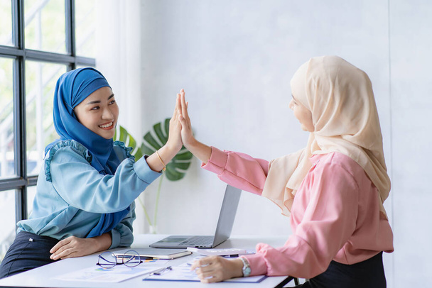 Twee mooie Aziatische moslimzakenvrouwen met hoofddoeken vieren hun prestaties door elkaars handen te klappen en hun handen op te steken.. - Foto, afbeelding