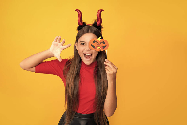 cheerful evil child in imp horns. happy halloween. devil kid with pumpkin party accessory. trick or treat. teen girl on yellow background. celebrate autumn party holiday. childhood fun. - Photo, Image