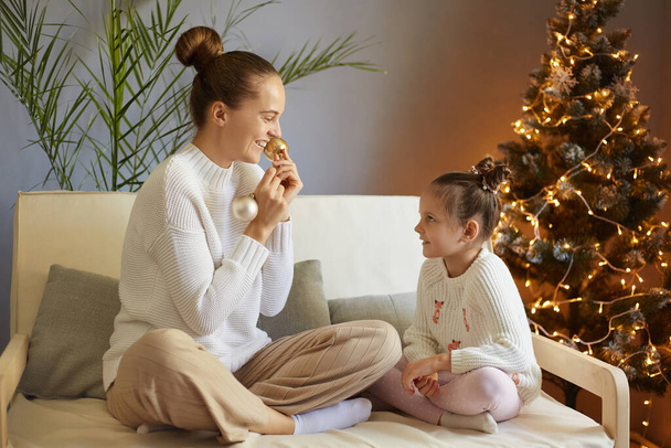 Winter Holidays. Portrait of cheerful woman and small girl in celebrating Xmas evening and New year at home, sitting on the couch playing with Christmas balls, laughing happily. - Foto, Imagem