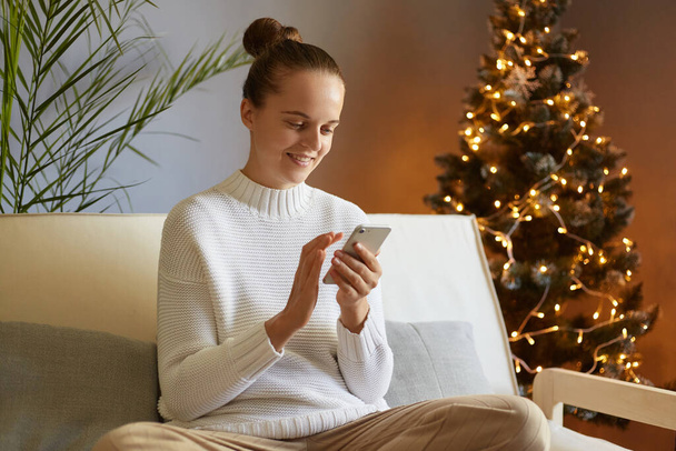 Portrait of pretty Caucasian woman with bun hairstyle sitting on cough using smart phone, chatting with her friends in social networks, celebrating christmas, posing near New year Eve. - Фото, зображення