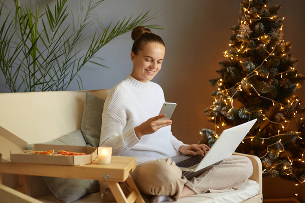 Indoor shot of woman using smartphone while working laptop at home on Christmas, sitting on sofa near fir xmas tree with lights, reading sms message on mobile during winter holidays. - Foto, Bild