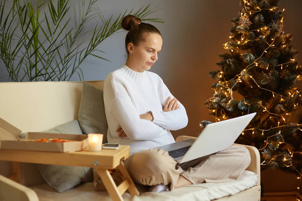 Sad angry woman working on laptop at home, female wearing white sweater sitting with crossed legs and folded arms, looking at notebook display with hate, works during Christmas holidays. - Foto, Bild