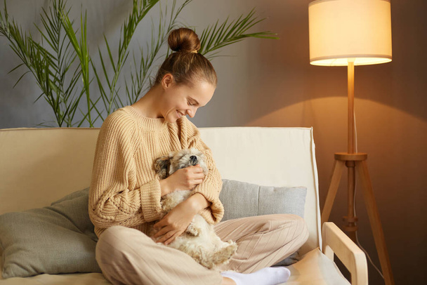 Young pretty woman in casual clothes hugging her beloved Pekingese dog sitting on the sofa in the living room of her cozy house, female smiling happily. - 写真・画像