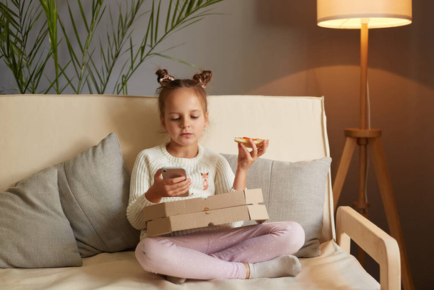 Horizontal shot of adorable cute charming little girl sitting at home on cough, relaxing at home while eating pizza, holding cell phone and watching funny video while eating fast food. - 写真・画像