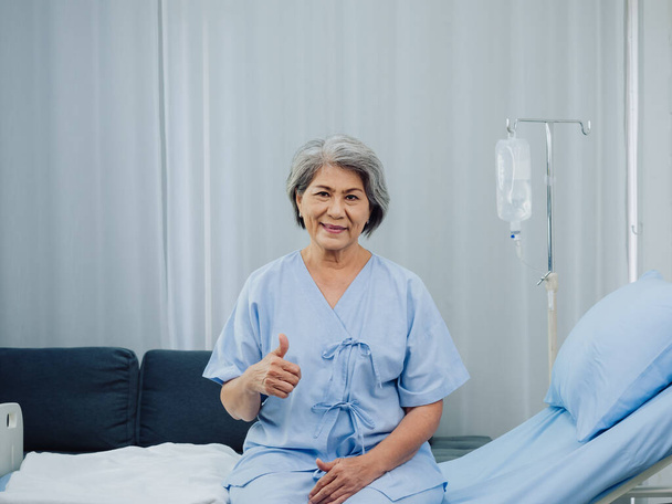 Onnellinen hymy Aasian vanhukset vanha naispotilas harmaat hiukset vaaleansininen mekko peukalo ylös istuessaan sängyssä lähellä lääketieteellistä IV napa seistä suolaliuoksella tippua valkoisessa huoneessa taustalla sairaalassa. - Valokuva, kuva