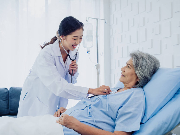 Beyaz takım elbiseli güzel bir Asyalı kadın doktor steteskop kullanarak hastanede yatan mavi elbiseli yaşlı bir kadının kalp atışlarını dinlerken gülümsedi.. - Fotoğraf, Görsel