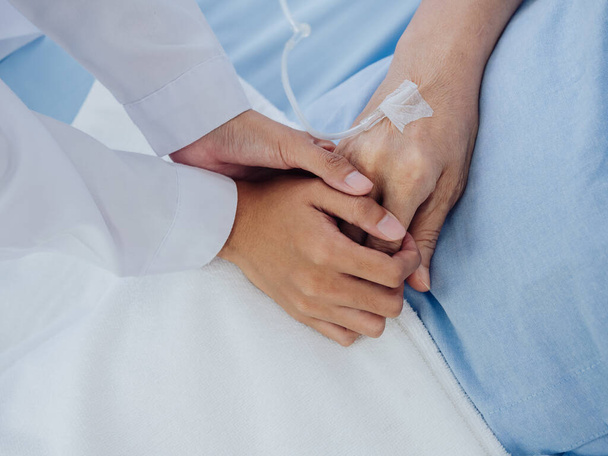 Close-up female doctor in white suit is holding on to elderly senior woman patient's hand in light blue dress with a saline hose lying on hospital bed, encouraging the patient concept. - 写真・画像