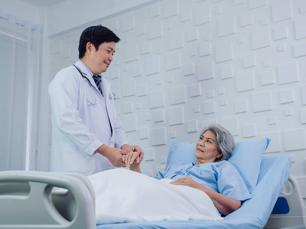 Rodzaj azjatyckiego lekarza w białym garniturze odwiedza, rozmawia i udziela wsparcia, trzymając za rękę szczęśliwą starszą kobietę w jasnoniebieskiej sukience leżącą na łóżku w pokoju szpitalnym. - Zdjęcie, obraz