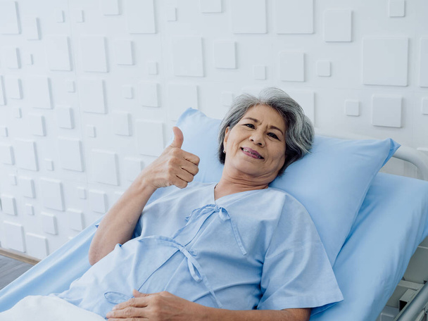 Χαρούμενη Ασιάτισσα ηλικιωμένη με γκρίζα μαλλιά, με τον αντίχειρα σηκωμένο και την κάμερα ξαπλωμένη στο κρεβάτι στο νοσοκομείο. Μια γριά με μπλε ρόμπα είναι άρρωστη στο κρεβάτι στο μοντέρνο λευκό δωμάτιο.. - Φωτογραφία, εικόνα