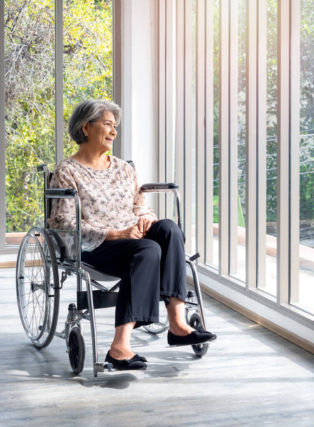 Happy Asian senior vrouw in casual jurk zit in een rolstoel, glimlachen en kijken uit het glazen raam met natuurlijk groen uitzicht, verticale stijl. Positief denken leidt tot een betere gezondheid bij ouderen. - Foto, afbeelding