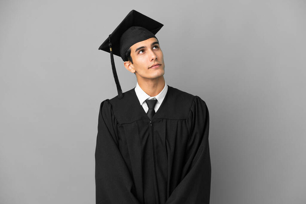 Νεαρός Αργεντινός απόφοιτος πανεπιστημίου απομονωμένος σε γκρίζο φόντο και κοιτώντας ψηλά - Φωτογραφία, εικόνα
