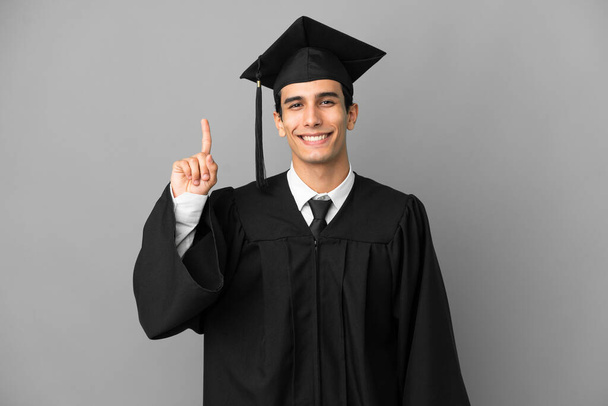 Νεαρός Αργεντινός απόφοιτος πανεπιστημίου απομονώνονται σε γκρι φόντο δείχνει και να άρει ένα δάχτυλο σε ένδειξη του καλύτερου - Φωτογραφία, εικόνα