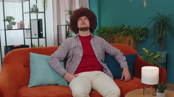 Repair work at neighbours concept. Irritated guy sits on couch cover ears with pillows annoyed by noisy neighbors suffer from headache wishes silence. Thin walls at home flat without sound insulation - Materiał filmowy, wideo