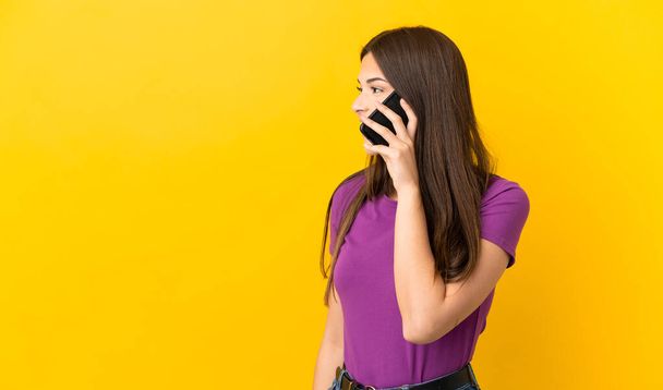 Teenager Brazilian girl over isolated yellow background keeping a conversation with the mobile phone with someone - Zdjęcie, obraz