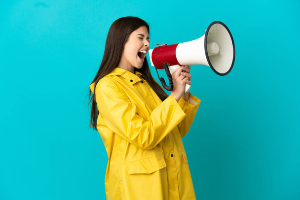 Teenager Brazilian girl wearing a rainproof coat over isolated blue background shouting through a megaphone - Photo, image