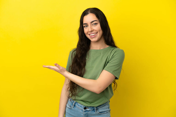 Młoda kobieta rasy kaukaskiej odizolowana na żółtym tle prezentująca pomysł, uśmiechnięta do - Zdjęcie, obraz