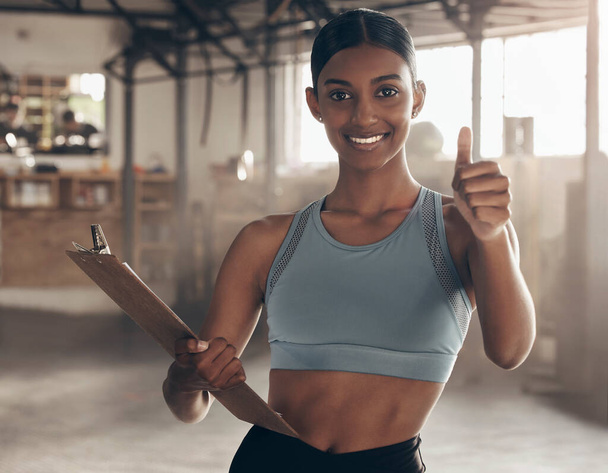 Our training facility is fully equipped and ready for you. Portrait of a sporty young woman showing thumbs up while holding a clipboard in a gym - Φωτογραφία, εικόνα
