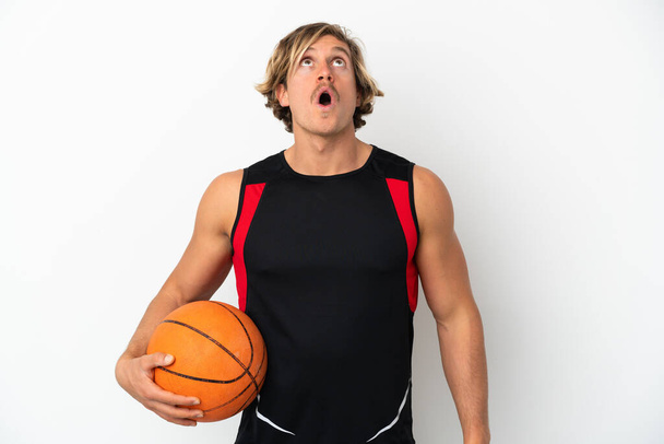 Jonge blonde man met een bal van basketbal geïsoleerd op witte achtergrond op zoek omhoog en met verrassende expressie - Foto, afbeelding