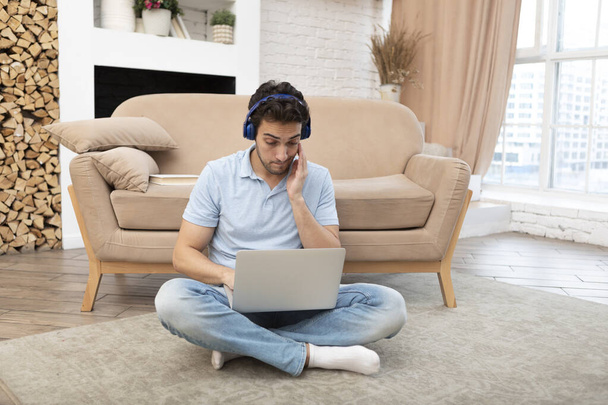 Fiatal férfi hallgató fülhallgatóban ül a padlón és laptopot néz. Online oktatás és távmunka - Fotó, kép