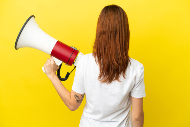 Redhead girl isolated on yellow background holding a megaphone and in back position - Photo, Image
