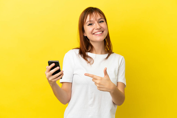 赤い頭の女の子は、携帯電話を使用して黄色の背景に隔離し、それを指す - 写真・画像