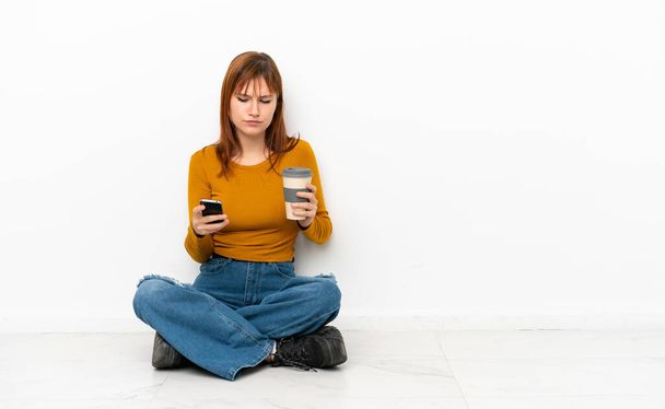 rousse fille assis sur le sol isolé sur fond blanc tenant café à emporter et un mobile - Photo, image