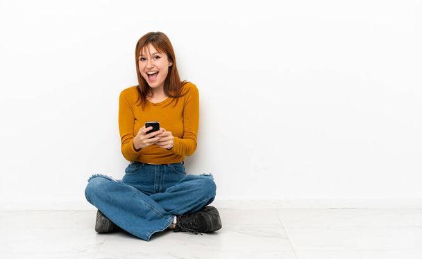 Ruda dziewczyna siedzi na podłodze odizolowany na białym tle zaskoczony i wysyłanie wiadomości - Zdjęcie, obraz