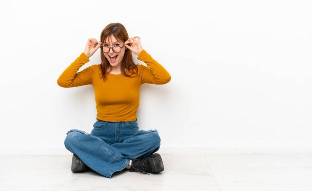 Ragazza rossa seduta sul pavimento isolata su sfondo bianco con occhiali e sorpresa - Foto, immagini