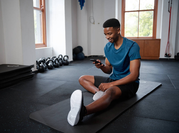 Young black man in sportswear on mobile phone while taking a break on exercise mat at the gym - Zdjęcie, obraz