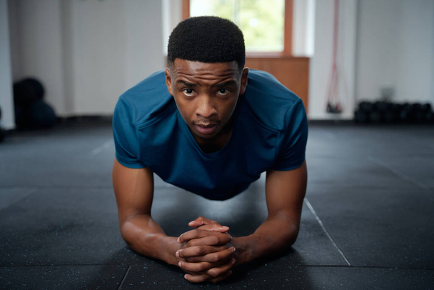 Focused young black man in sportswear doing plank exercises with hands clasped at the gym - Foto, Imagem