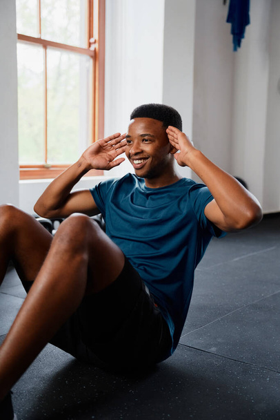 Confident young black man in sportswear smiling while doing sit-ups at the gym - Фото, изображение