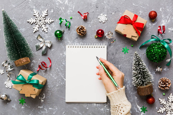 Γυναίκα που γράφει τα Χριστούγεννα για να κάνει λίστα στο σημειωματάριο σε χρωματιστό φόντο με χριστουγεννιάτικα στολίδια. - Φωτογραφία, εικόνα