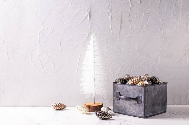 Christmas decorations. Metallic box with glass gold, black, silver decorative cones and white tree on white  textured background. Scandinavian minimalistic style. Still life. Place for text.  - Фото, изображение