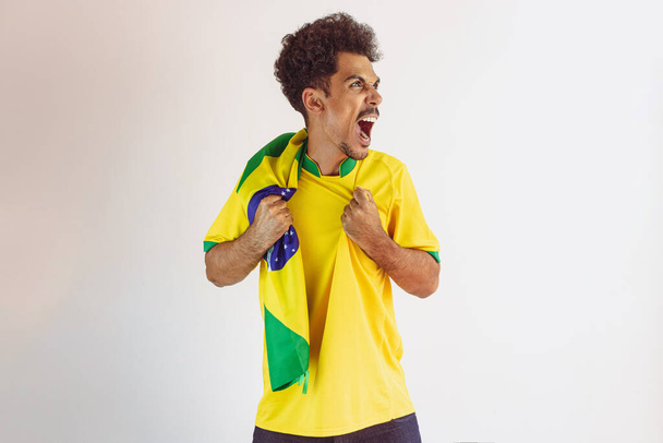 サッカーチームのシャツとブラジルの黒人男性ファンは白に隔離されました。カップを祝う旗を持つスポーツファン. - 写真・画像