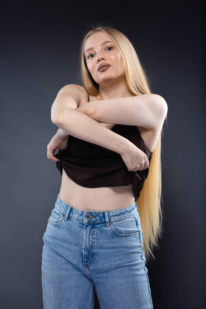 Young blonde woman in jeans and brown singlet on dark gray background - Photo, Image
