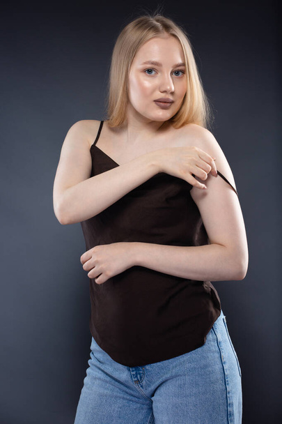 Young blonde woman in jeans and brown singlet on dark gray background - Foto, Imagen