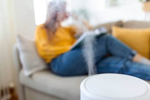 Очиститель воздуха в уютной белой гостиной для фильтра и очистки, удаляя пыль PM2.5 HEPA дома с женщиной, читающей книгу на диване, для свежего воздуха и здоровой жизни, Концепция загрязнения воздуха - Фото, изображение